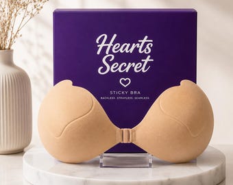 Puede incluir: Un sujetador adhesivo de color beige con un cierre frontal, colocado frente a una caja rectangular morada. La caja presenta el texto blanco Hearts Secret, un icono de corazón y las palabras STICKY BRA, BACKLESS, STRAPLESS, SEAMLESS.