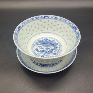 Könnte beinhalten: Eine weiße Porzellanschale mit passender Untertasse und blauen Unterglasurmustern. In der Mitte der Schale befindet sich ein detailliertes Drachenmotiv, während die Seiten und der Rand mit durchscheinenden Reiskornmustern und blauen geometrischen Bordüren verziert sind.