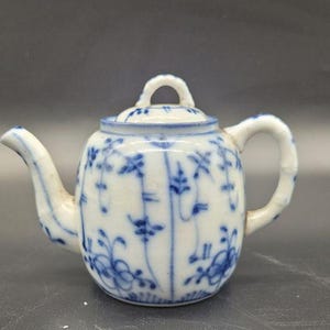 Puede incluir: Una pequeña tetera de cerámica blanca con un delicado patrón floral pintado a mano en azul. La tetera incluye una tapa a juego con un asa pequeña arqueada, un pico curvo y un mango texturizado que imita un tallo de bambú.