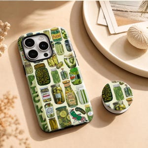 Puede incluir: Una funda para teléfono y un soporte circular a juego con un patrón repetitivo de varios frascos de vidrio llenos de pepinillos verdes. La funda incluye la palabra PICKLES impresa verticalmente en letras verdes grandes y un pepinillo.
