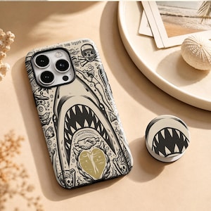 May include: A beige smartphone case and a matching circular phone grip featuring a black and white graphic illustration of a shark, a diver, a skull, and a handshake icon with the text Amity Means Friendship and a gold heart shape.