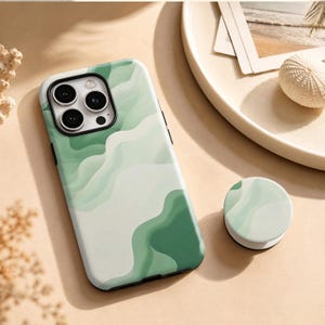Op de afbeelding: Een smartphonehoesje en een bijpassende ronde telefoonhouder met een abstract golvend patroon in diverse tinten salie-, munt- en bosgroen. Het hoesje heeft een rechthoekige uitsparing voor een camerasysteem met drie lenzen.