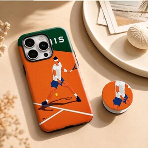 Può includere: Una custodia per smartphone e un supporto circolare abbinato con un'illustrazione grafica di un tennista su un campo in terra battuta arancione. Il design include una sezione superiore verde scuro con il testo bianco parziale IIS e una pallina.