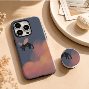 May include: A slate blue smartphone case and a matching circular phone grip. The case features a graphic of a black cat with small wings floating against a background of soft, painterly orange and pink clouds. The design is repeated on the grip.