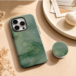 May include: A sage green smartphone case featuring a watercolor-style wash pattern with small white star-like accents and a matching circular collapsible phone grip. The case has a black camera module cutout with three circular lens openings.