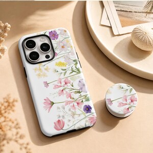 May include: A white smartphone case and a matching circular phone grip, both featuring a delicate watercolour-style floral pattern with pink, purple, yellow, and green wildflowers scattered across the surface against a clean white background.