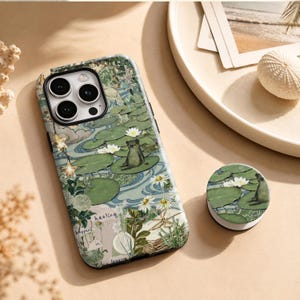 Könnte beinhalten: Eine Handyhülle und ein passender runder Handygriff mit einem Teichmotiv, grünen Seerosenblättern, weißen Seerosen, einem kleinen Frosch und goldenen Sternen. Das Design zeigt botanische Illustrationen und den Text "healing happens in".