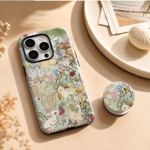 Peut inclure: Une coque de smartphone et un support de téléphone circulaire assorti présentant un fond vert clair avec une illustration détaillée d'animaux, dont un faon, des lapins et des canards, parmi des fleurs, des champignons et des papillons.