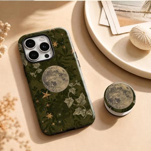 May include: A dark olive green phone case and a matching circular phone grip featuring a detailed full moon, scattered gold-toned stars, and light green ivy leaf patterns. The phone case has a cutout for a triple-lens camera system on the back.