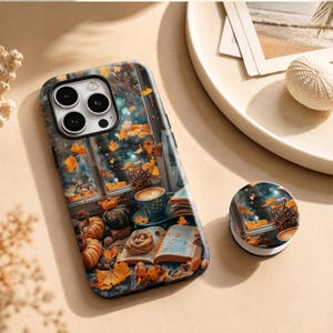May include: A smartphone case and a matching circular phone grip featuring a printed autumn scene. The design displays a window view with falling orange leaves, pumpkins, an open book, and a cup of coffee on a table, set against a dark background.