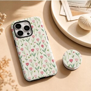 May include: A smartphone case and a matching circular phone grip featuring a cream-coloured background with a repeating pattern of pink tulips and green stems and leaves. The case has a square camera cutout with three circular lenses visible.