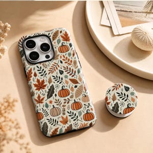 Op de afbeelding: Een smartphonehoesje en een bijpassende ronde telefoonhouder met een herhalend patroon van oranje pompoenen en diverse herfstbladeren in tinten bruin, beige en gedempt groen op een crèmekleurige achtergrond, met een zwarte uitsparing voor de camera.