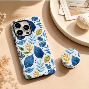 May include: A smartphone case and a matching circular phone grip featuring a white background with a repeating pattern of watercolour-style leaves in shades of deep blue, light blue, and yellow. The case has a cutout for a triple-lens camera.