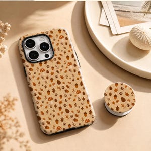 Op de afbeelding: Een smartphonehoesje en een bijpassende ronde telefoonhouder met een herhalend patroon van bruine en oranje paddenstoelen, eikels, dennenappels, blaadjes en kleine pootafdrukken op een lichtbruine achtergrond. De hoes heeft een uitsparing voor drie camera's.