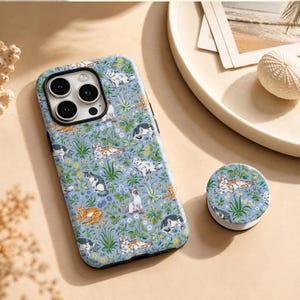 May include: A light blue smartphone case and a matching circular phone grip featuring a repeating pattern of various cats in different poses interspersed with small yellow flowers and green foliage. The case has a cutout for a triple camera system.
