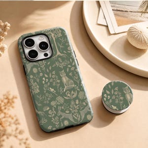 May include: A sage green smartphone case and a matching circular phone grip featuring a detailed white line art pattern of foxes, rabbits, hedgehogs, birds, trees, and various wildflowers scattered across the entire surface of both items.