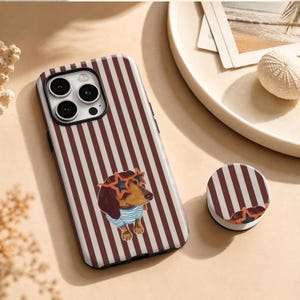 Pode incluir: Uma capa para smartphone e um suporte circular correspondente com um padrão de riscas verticais castanhas e brancas. A capa apresenta um cão dachshund castanho com óculos de sol cor de laranja em forma de estrela e uma t-shirt às riscas azuis e brancas.
