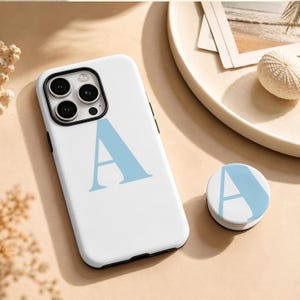 May include: A white smartphone case featuring a large, light blue letter A printed on the back, accompanied by a matching circular phone grip that also displays a light blue letter A on a white background, both designed for mobile devices.