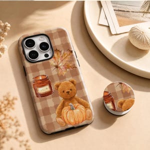 Op de afbeelding: Een telefoonhoesje en bijpassende ronde telefoonhouder met een bruin en beige ruitpatroon. Het ontwerp bevat een bruine teddybeer die een oranje pompoen vasthoudt, een brandende kaars in een glazen pot en een esdoornblad in oranje tinten.