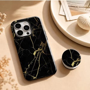 May include: A black smartphone case featuring a gold marble vein pattern and a matching circular collapsible phone grip. The case has a cutout for a triple-lens camera system on the back, showcasing a sleek, dark surface with metallic gold accents.