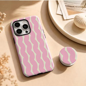 Puede incluir: Una funda rectangular para smartphone y un soporte circular a juego con un patrón repetitivo de rayas rosas verticales y onduladas sobre un fondo amarillo pálido. La funda tiene un marco negro para el sistema de cámara de triple lente.