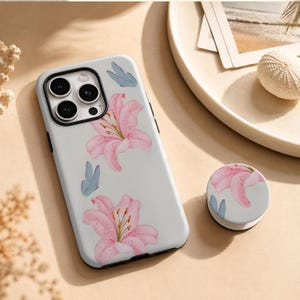 May include: A white smartphone case and a matching circular phone grip featuring a watercolour print of pink lily flowers and small light blue butterflies. The case has a black camera lens frame and cutouts for the triple camera system and flash.