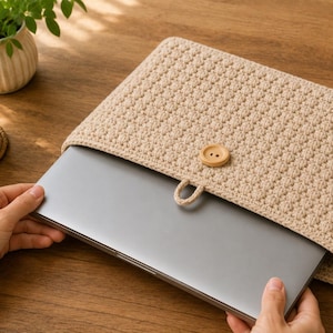 May include: A beige crochet laptop sleeve featuring a textured stitch pattern and a single wooden button closure with a matching loop. The sleeve is rectangular and designed to hold a flat electronic device, showcasing a neutral color palette.