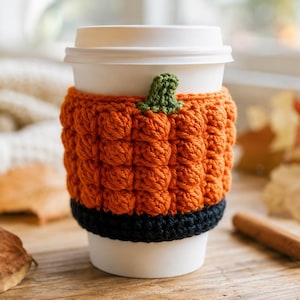 May include: A white disposable coffee cup featuring a textured orange crochet cozy designed to resemble a pumpkin, complete with a small green stem at the top and a solid black crochet band at the base, providing a decorative sleeve for the cup.