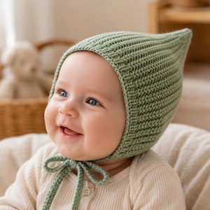 Peut inclure: Un bonnet tricoté vert sauge avec un sommet pointu et une texture côtelée. Le bonnet présente une mentonnière avec un nœud, fabriqué dans une matière en laine douce qui couvre la tête et les oreilles, offrant un ajustement serré pour un bébé.