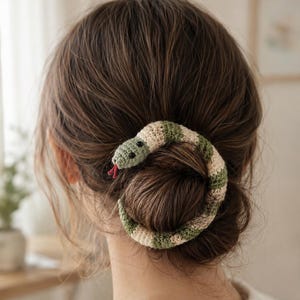 Peut inclure: Un accessoire de coiffure en crochet en forme de serpent avec des bandes alternées de fil crème et vert sauge. Le serpent présente de petits yeux en perles noires et une fine langue en fil rouge, enroulé autour d'un chignon pour maintenir les cheveux.