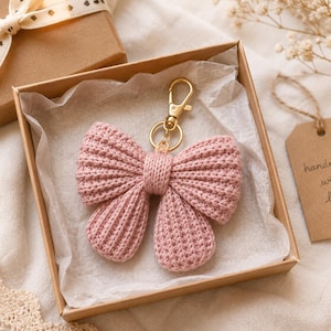 May include: A dusty pink crocheted bow keychain featuring a gold-toned metal lobster clasp and ring. The bow is crafted with a ribbed texture and is presented inside a square cardboard gift box lined with white tissue paper.