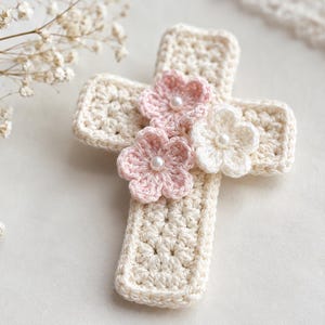 May include: A cream-coloured crochet cross featuring three small, layered flowers attached to the centre. Two of the flowers are light pink and one is white, each accented with a small white pearl bead at the centre of the floral arrangement.
