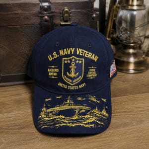 May include: A navy blue baseball cap featuring gold embroidery. The front displays U.S. NAVY VETERAN, an anchor shield, and the text ANCHORS AWEIGH and COURAGE COURAGE COMMITMENT. The brim shows a gold embroidered aircraft carrier and helicopters.