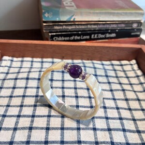 May include: A circular bracelet constructed from rectangular segments of iridescent white mother-of-pearl. The center features a large, spherical purple amethyst bead flanked by two silver-toned metal spacers embedded with small, clear, and purple crystals.
