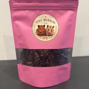 Puede incluir: Una bolsa rosa con cierre y una ventana transparente en la parte inferior que contiene virutas de madera de color marrón oscuro. La etiqueta presenta un logotipo circular con dos hámsteres y el texto Tiny Burrow Co. sobre la ventana.