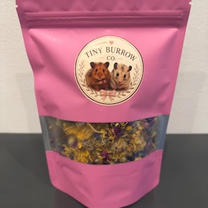 Op de afbeelding: Een roze stazak met een doorzichtig venster dat een mix van gedroogde bloemen laat zien. De voorzijde heeft een rond etiket met de tekst Tiny Burrow Co. en een illustratie van twee hamsters omringd door een krans en een kleine strik.