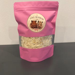 Puede incluir: Una bolsa rosa con base plana que contiene material de lecho triturado de color claro. El frente presenta una etiqueta circular con el texto Tiny Burrow Co. y una ilustración de dos hámsteres, uno marrón y otro blanco, en un marco decorativo.