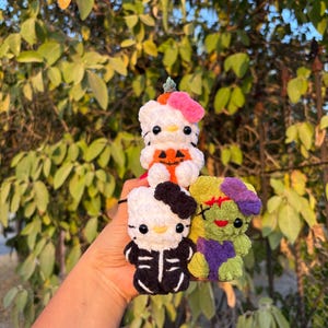 Puede incluir: Tres pequeñas figuras de peluche tejidas a ganchillo que incluyen un gato blanco con un lazo rosa y una calabaza naranja, un gato blanco con un disfraz de esqueleto negro y un monstruo verde con detalles morados y una cicatriz roja cosida.