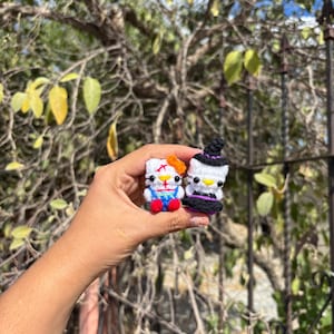 Puede incluir: Dos pequeñas figuras de ganchillo, una que representa un gato blanco con un lazo naranja, peto azul y zapatos rojos, y la otra un gato blanco con un sombrero de bruja negro y un vestido morado, ambos con ojos negros y nariz amarilla.