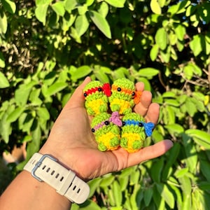 Puede incluir: Cuatro pequeñas figuras de ganchillo hechas a mano con forma de tortuga, cada una con una máscara de color distinto en rojo, naranja, morado y azul. Las figuras tienen cuerpos de color verde claro, vientres amarillos y ojos de cuentas negras.
