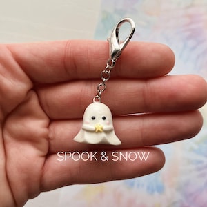 May include: A white ghost charm with black eyes, pink cheeks, and a small yellow star held in its hands. The charm is attached to a silver-toned metal lobster clasp by a short chain. The text SPOOK & SNOW is printed in white at the bottom.