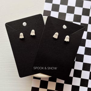 Op de afbeelding: Twee paar kleine, witte oorknopjes in de vorm van een spookje met zwarte ogen en mondjes. Elk paar is bevestigd op een rechthoekige zwarte displaykaart met de witte tekst SPOOK & SNOW onderaan en een rond gat aan de bovenzijde.