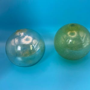 May include: Two spherical glass floats, one with a reflective silver finish and the other with a translucent light green tint. Both items feature a small circular opening at the top and a smooth, rounded surface with visible air bubbles in the glass.