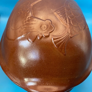 May include: A smooth, brown, dome-shaped ceramic mold featuring an embossed design of a rooster and a fish. The surface has a glossy finish, and the object is rounded with a wide base, showcasing detailed relief carvings of the animal figures.