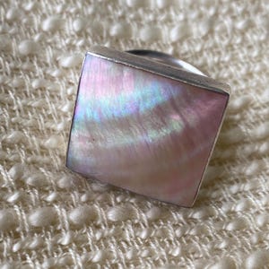 May include: A square ring featuring a flat, iridescent mother-of-pearl inlay with soft pink and blue hues, set within a polished silver-toned metal frame. The surface of the inlay displays a natural, swirling pearlescent texture.