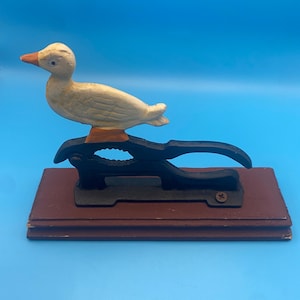 May include: A cast iron nutcracker featuring a white duck with an orange beak mounted on a black metal lever mechanism. The entire assembly is secured to a rectangular brown wooden base with a visible screw on the right side of the black frame.