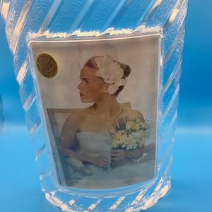 May include: A clear glass picture frame with a diagonal ribbed texture and a smooth central rectangular panel. A gold circular sticker with the text Cristal d'Arques France is attached to the upper left corner of the frame, which holds a photograph.