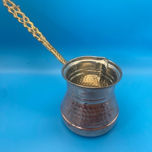 May include: A copper coffee pot with a hammered texture and a wide, rounded base. The pot features a long, ornate, gold-toned metal handle with a lattice-like cutout design and a wide, open top with a small pouring spout on the rim.