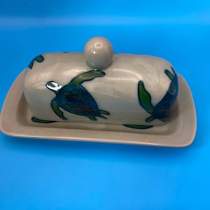 May include: A rectangular ceramic butter dish with a matching lid featuring a rounded knob handle. The off-white surface is decorated with hand-painted green and blue sea turtles swimming in various directions across the lid and base.
