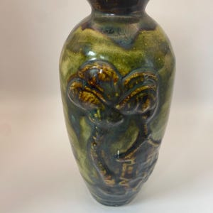 May include: A ceramic vase with a glossy finish in shades of olive green, brown, and dark blue. The body of the vase features a raised, embossed relief design depicting a palm tree and a small structure, with a narrow neck and flared rim.